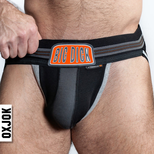 A person wearing a black jockstrap with a prominent orange waistband branded 'BIG DICK', showing a 3-panel pouch design and slider-strap butt-straps.